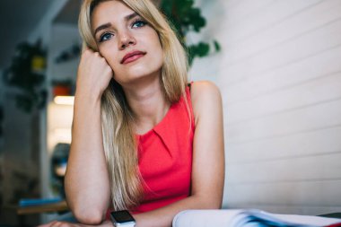 Düşünceli sarışın kız öğrenci kitaplarla masada oturuyor ve kafeteryada çalışırken tatilin hayalini kuruyor ve ödev yapıyor.