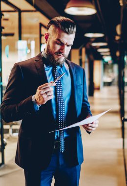 Resmi giyinmiş, kuşkucu sakallı girişimci modern ofis içinde ayakta duran bir iş sözleşmesi giyiyor. Düşünceli erkek gururlu CEO elinde kalemle ofiste mali raporunu kontrol ediyor.
