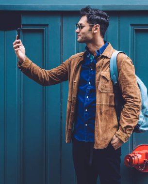 Sırt çantalı, modaya uygun giysileri ve güneş gözlükleriyle gülümseyen genç adam New York caddesinin mavi binasında dururken selfie çekiyor.
