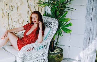 Kırmızı elbiseli, rujlu, beyaz rahat sandalyeli, yeşil tropikal bitkilerin ve yontulmuş duvarların arkasındaki kameraya bakan modern kızıl kadın.