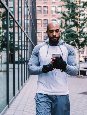 Ciddi, kel, sakallı, spor giyimli Amerikan erkek. Akıllı telefon kullanıyor ve modern binanın yanında sokak ortasında müzik dinliyor.
