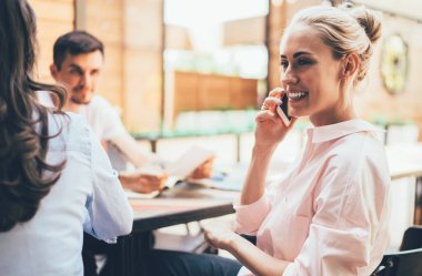 Neşeli yetişkin girişimci, açık modern açık uzay ofisindeki iş arkadaşlarıyla toplantı sırasında masasında otururken ve gözlerini kaçırırken telefonla konuşuyor.