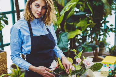 Çiçek hobisi için harcadığı süre boyunca çiçekçilikte bahçeyle uğraşan pozitif botanikçinin yarım boy portresi. Profesyonel önlüklü başarılı bir kadın kendi konservatuarında dikilirken kameraya bakıyor.