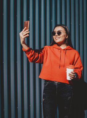 Pozitif Çinli kadın blogcu şık güneş gözlüklü kahve molası sırasında kenar mahallelerde fotoğraf çekiyor mutlu gülümseyen hippi kız cep telefonuyla yeni görüntüler yaratmak için zaman harcıyor.