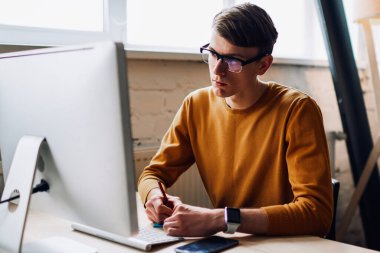 Gözlüklü genç adam, modern bilgisayarda internet üzerinden webinar öğrenirken ofisindeki masaüstünde oturduğu için not defterine notlar yazarak göz koruması sağlıyor.