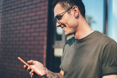 Gözlüklü, sıradan giyinen ve şehirdeki modern binalara karşı sokakta akıllı telefonuyla etkileşim kuran yakışıklı, mutlu bin yıllık adam.