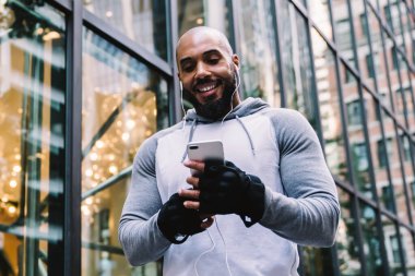 Gülümseyen Afrikalı Amerikalı bir adamın spor giyim kuşamında müzikten zevk alması ve cam ofis binasının yanında dikilirken akıllı telefon kullanması.