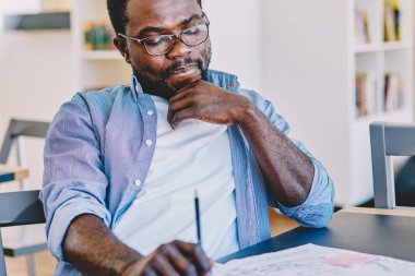 Sıradan giyinmiş, gözlüklü zeki Afro-Amerikan erkek. Masada oturup ofiste proje üzerinde çalışırken planlar üzerinde kafa yoruyor.