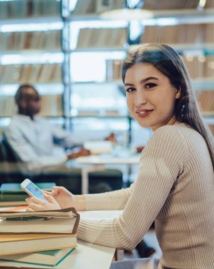 Kütüphanede oturmuş kameraya gülümserken resmi giyinmiş, akıllı telefonlara göz atan mutlu kadının yan görüntüsü.