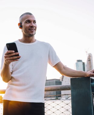 Beyaz gömlekli, kulaklıklı, akıllı telefonlu, metalik çit ve nehrin yanında duran kısa saçlı genç adam şehrin arka planına yaslanmış.