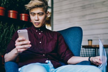 Modern akıllı telefondan 4G internete bağlı takipçilerden gelen tebligatı okuyan düşünceli Çinli hipster blogcusu modern uzay sandalyesinde oturuyor. Japon genç adam cep telefonuyla video izliyor.
