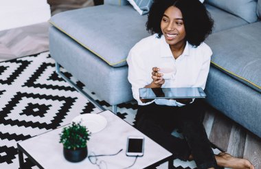 Kahvesini ve dijital dokunmatik defterini elinde tutarak gülen koyu tenli hippi blogcu modern bir dairede şık bir iç dekorasyonla oturuyor. Neşeli, düşünceli, Afro-Amerikan genç kadın.