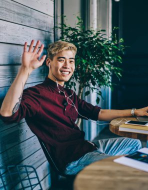 Uzaktan kumanda sırasında modern laptopa bağlı kulaklıkla müzik dinlerken el sallayan mutlu Çinli hipsterin portresi. İnternetteki yazılımı güncelleyen olumlu Japon genç adam.