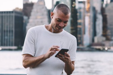 Genç gülümseyen etnik adam kulaklıkla müzik dinlerken akıllı telefon kullanıyor ve nehir ve şehrin bulanık arka planına karşı korkulukların yanında duruyor.