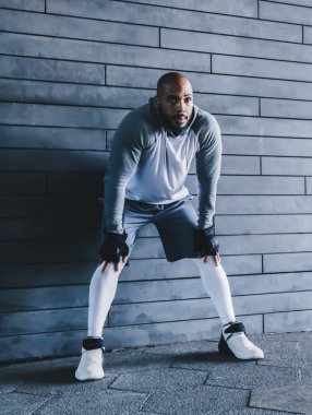 Gri spor giyimli, kel, yorgun ve sakallı bir adam. Duvara yaslanmış siyah eldivenleri var. Elleri dizlerinin üstünde ve dinlenirken başka yere bakıyor.