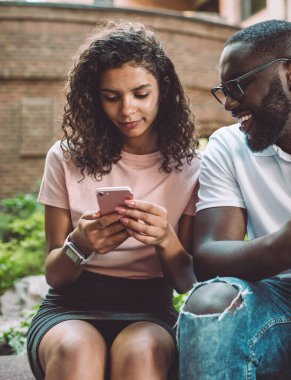 Gözlüklü, gülümseyen hippi, akıllı telefon ekranına bakarak sevgili kız arkadaşının yazdığı SMS mesajlarını kontrol ediyor. Aşık çift akıllı telefon cihazlarında 4G kablosuz internet kullanıyor.