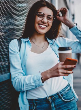 Gözlüklü neşeli bir genç kadının portresi kameraya gülümseyen nefis bir kahve tutarken ve şehir ortamında akıllı telefonu elinde tutarken. Modern hücresi olan pozitif hipster kız caddede duruyor.