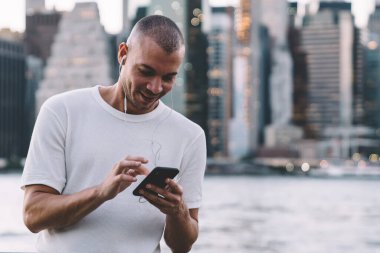 Genç gülümseyen etnik adam kulaklıkla müzik dinlerken akıllı telefon kullanıyor ve nehir ve şehrin bulanık arka planına karşı korkulukların yanında duruyor.