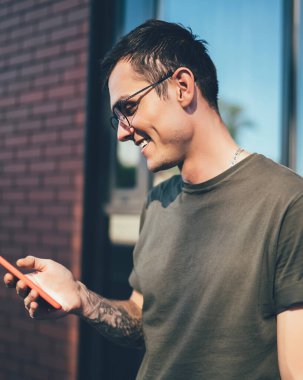 Gözlüklü, sıradan giyinen ve şehirdeki modern binalara karşı sokakta akıllı telefonuyla etkileşim kuran yakışıklı, mutlu bin yıllık adam.