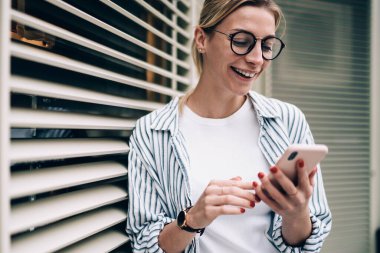 Neşeli, genç, güzel bir bayan günlük kıyafetler ve moda gözlükler takıp gülüyor, akıllı telefonlara bakıyor ve bina yakınlarında komik videolar seyrediyor.