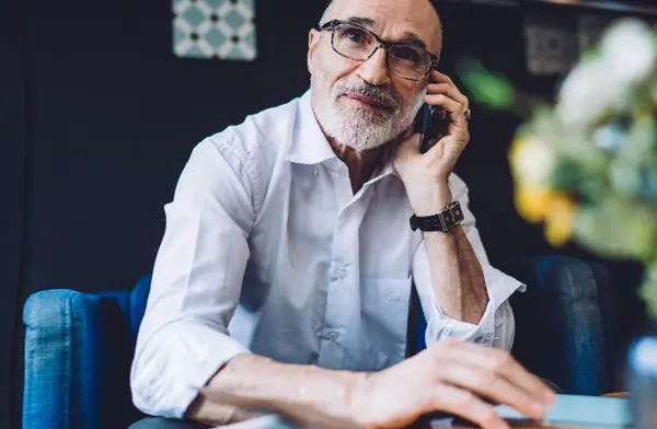 Bald gray bearded male with eyeglasses in white shirt sitting at table on blue chair and looking away while speaking on phone