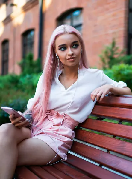 Slim lady in white shirt and pink shorts with long pink hair listening to music on mobile phone sitting on wooden bench in street