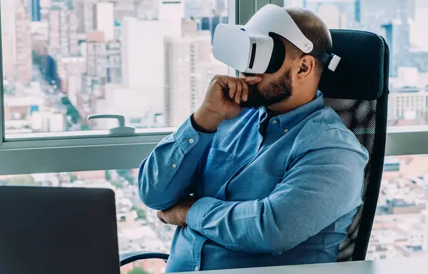 Man wearing glasses of virtual reality while sitting at computer table in contemporary office with cityscape behind window on background