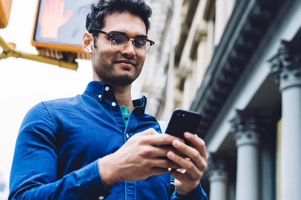 Yakışıklı, tişörtlü ve gözlüklü yetişkin bir adam. Gün ışığında kavşakta dikilirken akıllı telefondan mesaj atıyor.