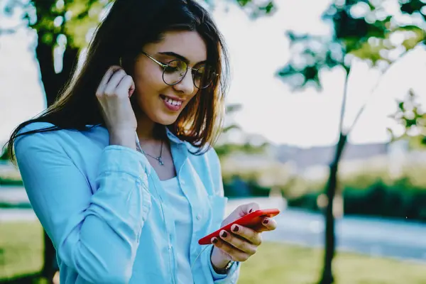 Smiling young woman listening audio in earphones from music app on modern smartphone device standing in outdoor in park.Positive hipster girl enjoying playlist on digital mobile phone in urban setting