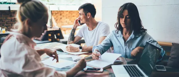 stock image Friends sitting at table with notebooks and laptop with blank screen while focused young women discussing written plan and man talking on smartphone