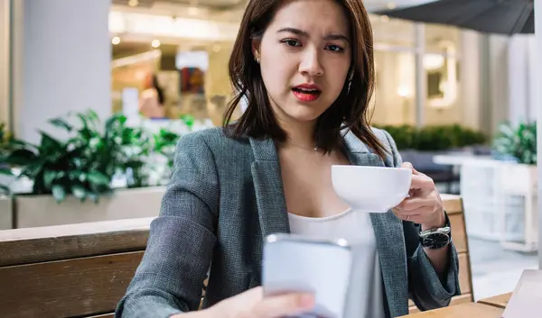 Young Asian confused woman with short hair and opened mouth holding coffee cup sitting at desk and having problems with smartphone in cafe