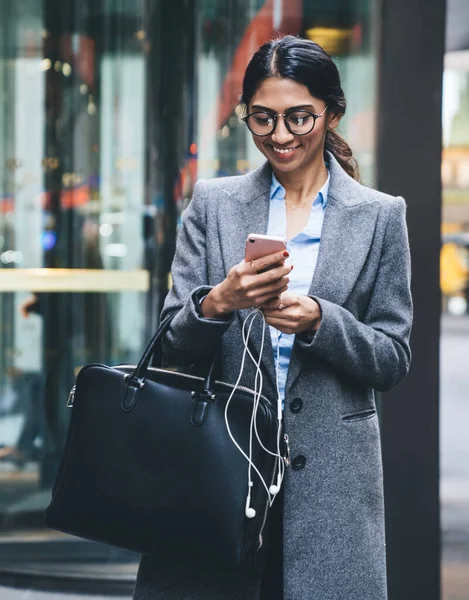 Smiling business lady in elegant outfit and eyeglasses holding leather bag surfing mobile while walking on street of New York city