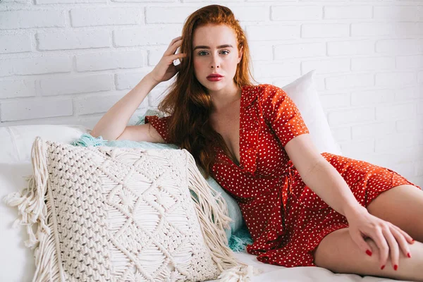 Woman with long red hair lying on white bed near pillows and leaning on hand while looking at camera at home
