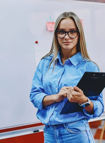 Portrait of charming young woman in eyeglasses working coach holding folder and standing against flip chat in classroom.Attractive female teacher looking at camera before workshop in modern office