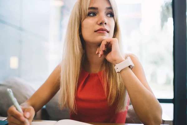 Young blond female in casual outfit looking away and thinking while sitting at table in cozy cafe and making notes in open planner
