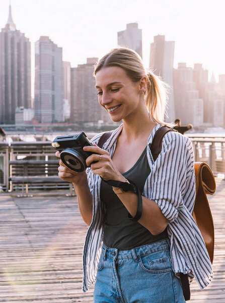 Cheerful tourist enjoying time for recalls to pleasant moments while looking at pictures on instant camera, happy woman with vintage equipment smiling at urban setting with Manhattan on background