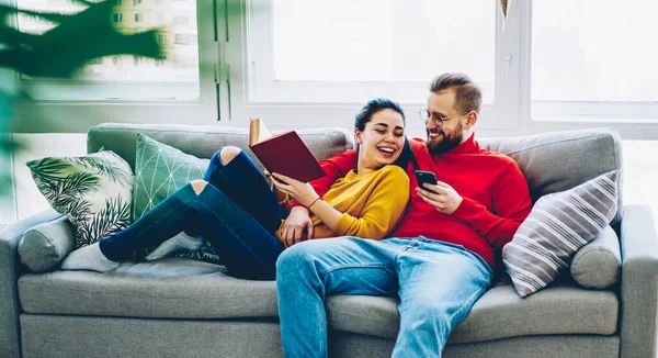 Happy young family having perfect relationships resting together on sofa in living room joking and communicating, romantic couple recreating with technology and book having fun on free time at home