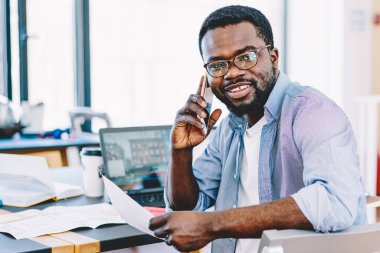 Gülümseyen Afro-Amerikan erkek cep telefonuyla konuşuyor. Elinde bilgisayarla masada oturuyor.