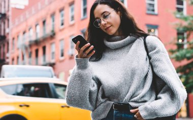 Gözlüklü, siyah çantalı, kot ve kazaklı çekici bir kadın. Şehir caddesinde faiziyle akıllı telefon kullanıyor.