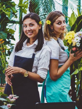 Başarılı bahçıvanların yarım boy portresi gündelik önlükler giyip ellerinde taze çiçekler tutan ve sıcak evde çalışırken kameraya bakan ortaklar işbirliği kavramı.