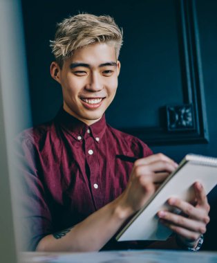 Modern dizüstü bilgisayarda Webinar 'ı izlerken notlar alan neşeli Çinli genç adam. Modern uzayda oturan notlara resim çizen pozitif Japon hipster öğrenci.