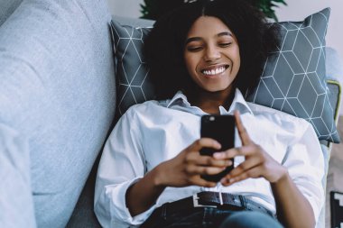 Koyu tenli neşeli hipster blogcu evde 4G internet kullanarak internette sohbet ederken gülüyor. Kanepede dinlenen ve arkadaşlarıyla mesajlaşan Afro-Amerikan genç kadın.