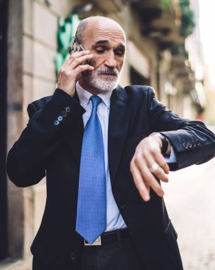 Resmi kıyafetli, beyaz sakallı, kendine güveni tam bir işadamı caddede saati kontrol ederken bulanık bina ve ağaçların arkasındaki kol saatine bakıyor.