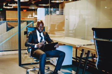 Resmi giyinmiş Afrikalı Amerikalı işadamı modern akıllı telefondan sohbet ediyor ve dijital tablette çalışıyor. Ofiste kablosuz internet kullanıyor. Koyu tenli girişimci profilini güncelliyor.