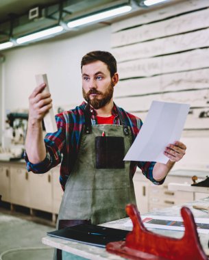 Önlüklü sakallı erkek, çalışma masasının yanında dururken küçük tahta tahtaları inceliyor ve marangoz atölyesindeki taslak ile ayrıntıları karşılaştırıyor.