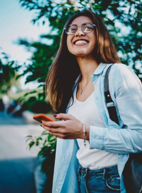 Yeşil parkta sırt çantalı pozitif hippi kız eğleniyor. Elinde dijital cep telefonu olan neşeli beyaz kadın şehir ortamında havanın ve havanın tadını çıkarıyor.