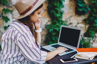Moda şapkalı genç beyaz kadın dışarıda kafede vakit geçirirken dizüstü bilgisayardan video seyrediyor, hipster kız sosyal ağlardan ve bloglardan haber okuyor ve netbook ve wifi bağlantısı kullanıyor.