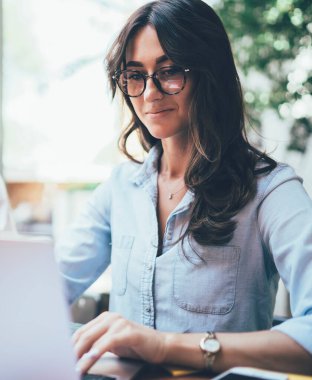 Gözlüklü ve mavi tişörtlü düşünceli yetişkin bir kadının güneşli bir günde terasta iş arkadaşlarıyla masasında otururken ekrana odaklanması ve dizüstü bilgisayarla etkileşime girmesi.
