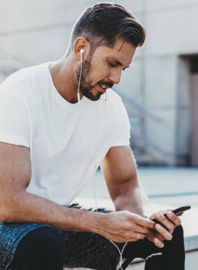 Spor kıyafetleri giymiş erkek sporcu dışarıda kardiyo eğitiminden sonra dinleniyor. Akıllı telefon zımbırtısıyla e-postaları kontrol ediyor ve koşmak için müzik listesi seçiyor. Elektronik kulaklıklı adam cep telefonu kullanıyor.