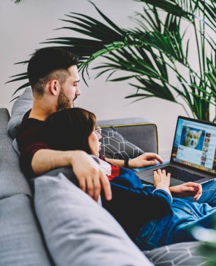 Genç bir adam ve bir kadın, rahat oturma odasında rahat bir kanepe, ilişki ve iletişim konsepti üzerinde dinlenirken ücretsiz ev Wi-Fi 'sine bağlı sosyal ağda sohbet etmek için internette geziniyorlar.